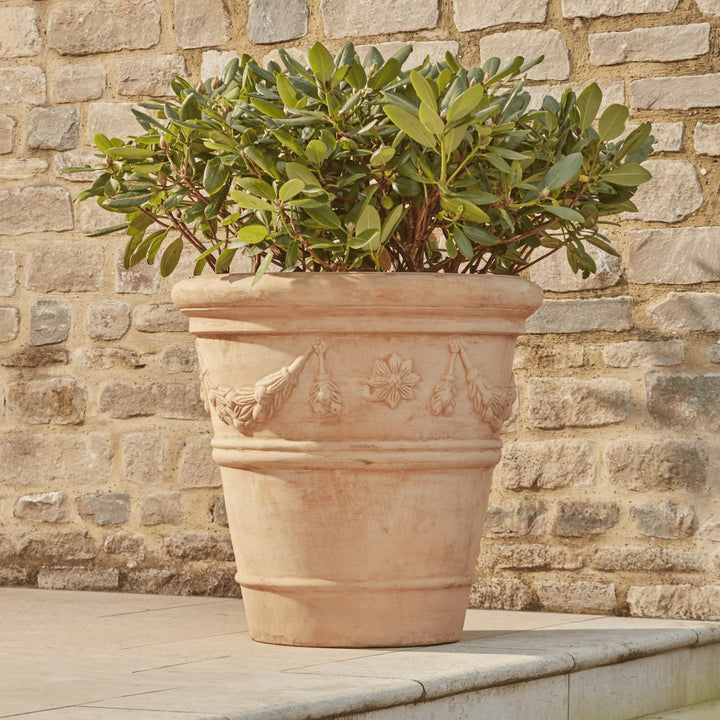 Large terracotta pot with a delicate floral garland design, hand-finished with a slightly weathered appearance. The pot has a softly tapered shape, a wide rim, and is placed outdoors with plants growing inside, against a stone wall backdrop.