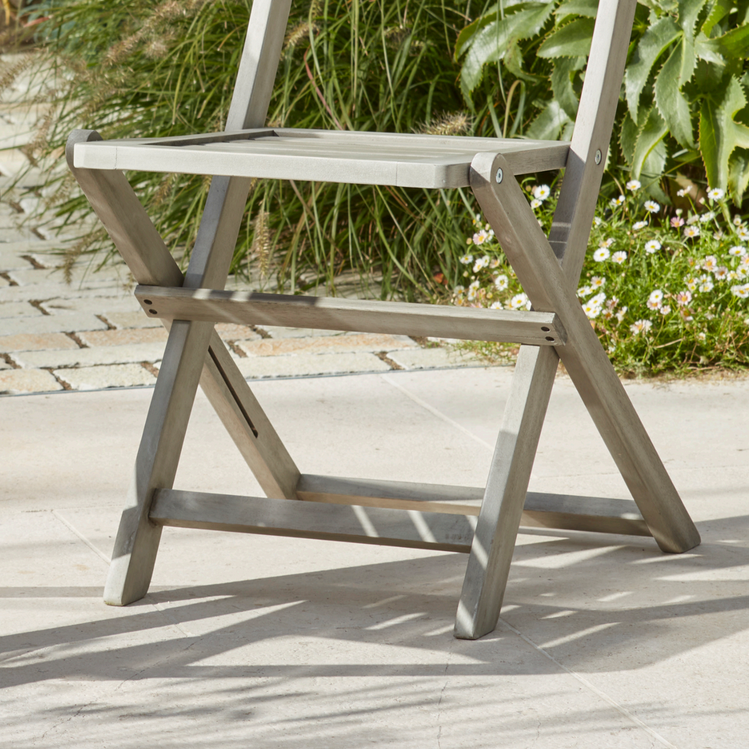 Close-up of the Repton Wooden Bistro Chair, highlighting its sturdy foldable frame and weathered grey finish. The slatted seat and cross-leg design offer both style and functionality, making it an ideal choice for outdoor patio, garden, or balcony seating. Positioned on a stone patio with lush greenery in the background.