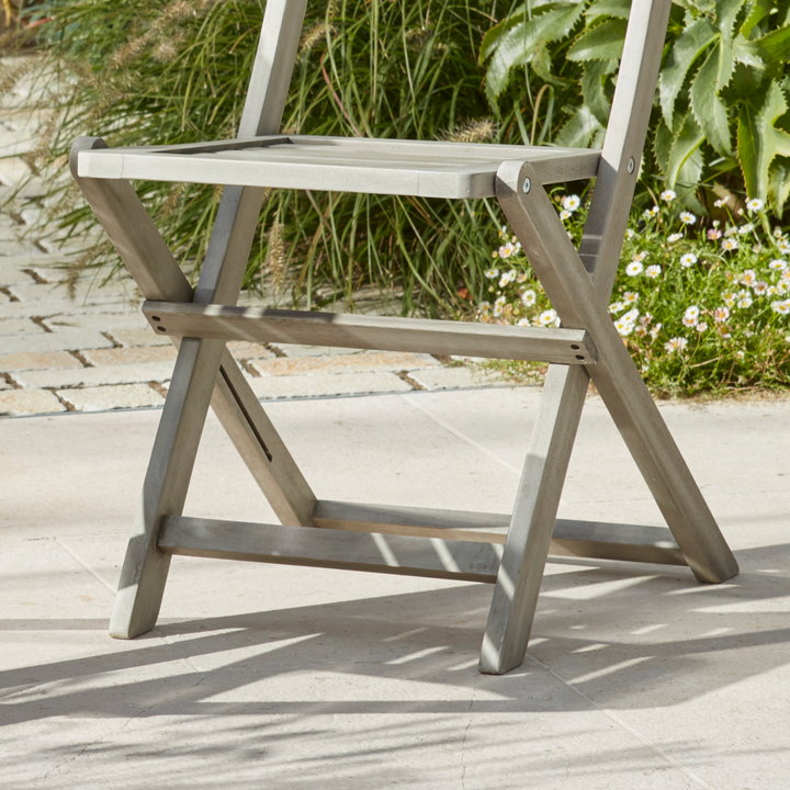 Close-up of the Repton Wooden Bistro Chair, highlighting its sturdy foldable frame and weathered grey finish. The slatted seat and cross-leg design offer both style and functionality, making it an ideal choice for outdoor patio, garden, or balcony seating. Positioned on a stone patio with lush greenery in the background.