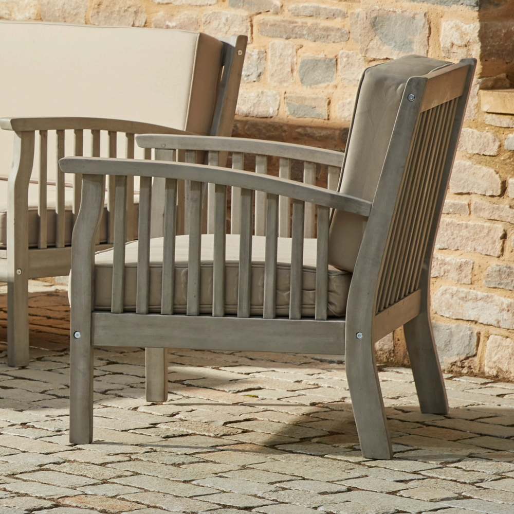 Close-up of the Repton Wooden Lounge Armchair, featuring a slatted backrest, curved armrests, and a weathered grey finish. The chair includes a plush beige cushion for added comfort, making it an elegant and durable choice for outdoor patio and garden seating. Positioned against a rustic brick wall on a stone-paved surface.