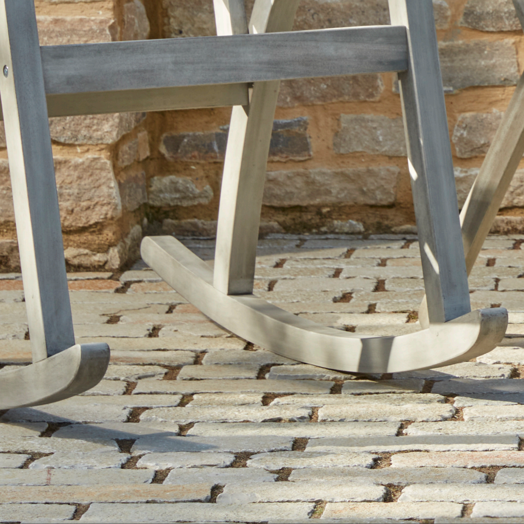 Close-up of the Repton Wooden Rocking Chair’s curved rockers, showcasing its sturdy construction and weathered grey finish. Designed for smooth, gentle movement, this outdoor rocking chair is perfect for relaxing on patios, porches, and garden spaces. Set on a rustic stone-paved surface against a brick wall.