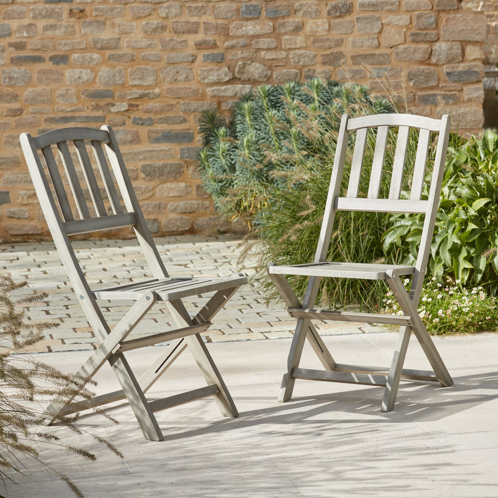 A pair of Repton Wooden Bistro Chairs with a weathered grey finish, elegantly positioned on a stone patio. These foldable wooden garden chairs feature a slatted backrest and seat, offering a classic yet functional outdoor seating solution. Set against a rustic brick wall with lush greenery, perfect for patios, balconies, and garden spaces.