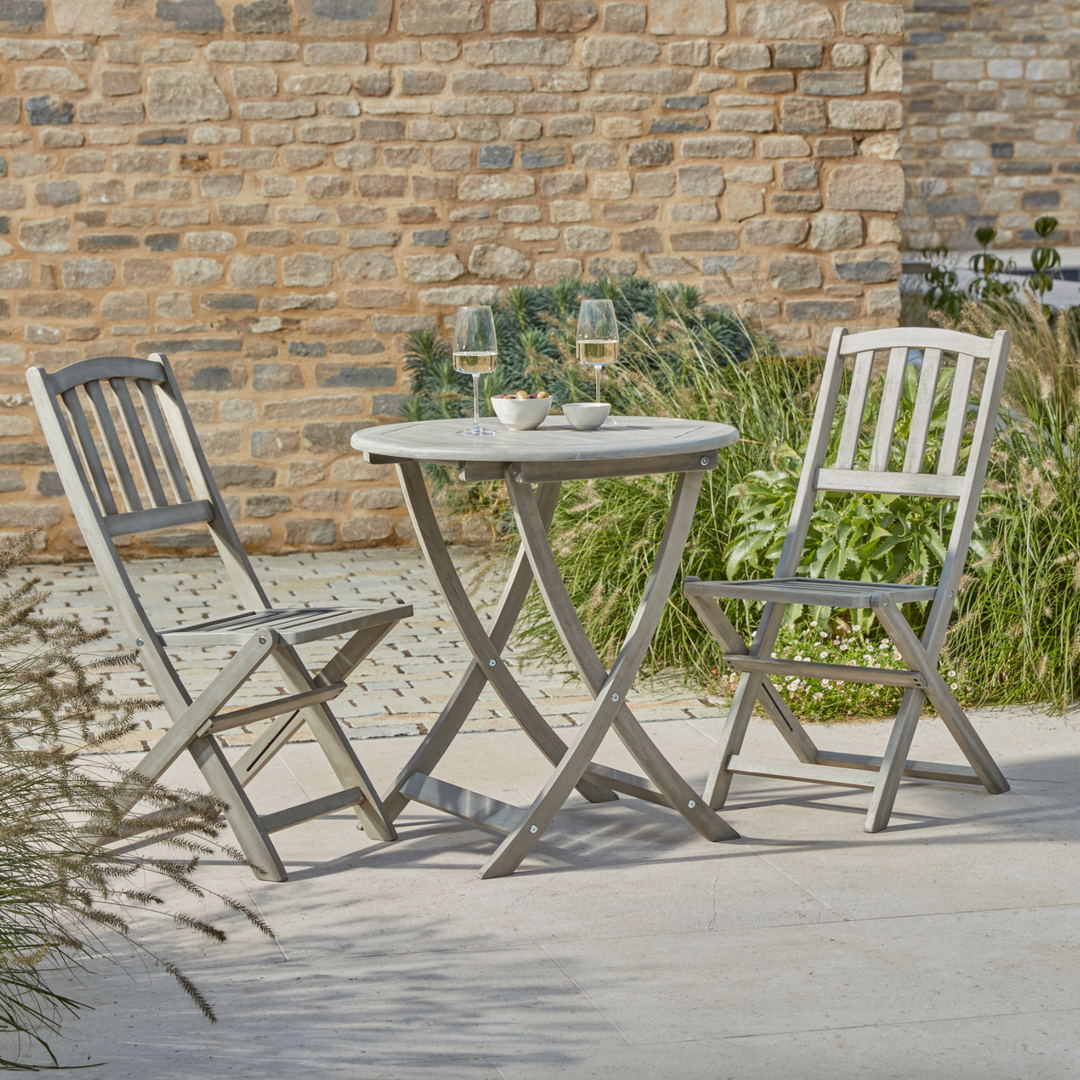 The Repton Wooden Bistro Set, featuring a round table and two folding chairs in a weathered grey finish, is elegantly arranged on a stone patio. The setup includes two wine glasses and small bowls of snacks, creating a charming outdoor dining scene against a rustic brick wall and lush greenery.