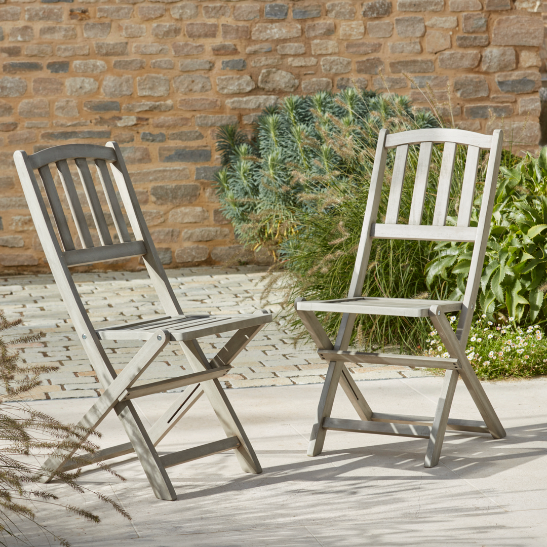 A pair of Repton Wooden Bistro Chairs with a weathered grey finish, elegantly positioned on a stone patio. These foldable wooden garden chairs feature a slatted backrest and seat, offering a classic yet functional outdoor seating solution. Set against a rustic brick wall with lush greenery, perfect for patios, balconies, and garden spaces.