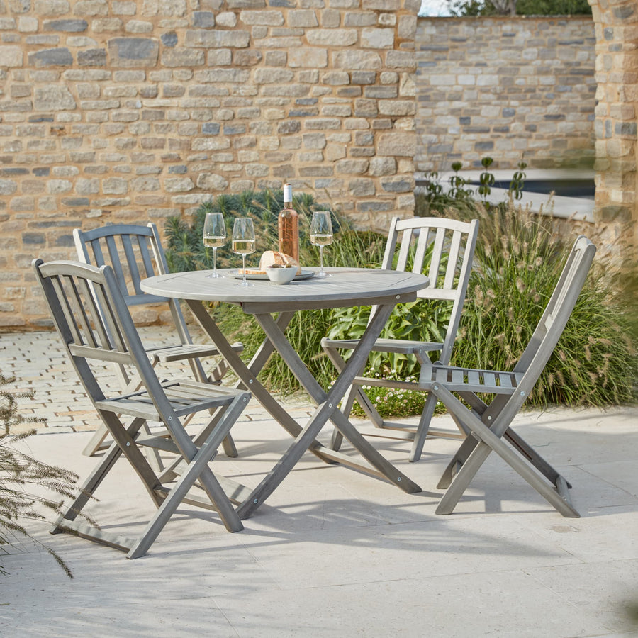 The Repton Wooden Dining Set, featuring a round foldable table and four matching wooden bistro chairs in a weathered grey finish. Styled on a stone patio with a bottle of wine, glasses, and bread, this outdoor dining set is perfect for garden gatherings. Set against a rustic brick wall with lush greenery for a charming alfresco dining experience.