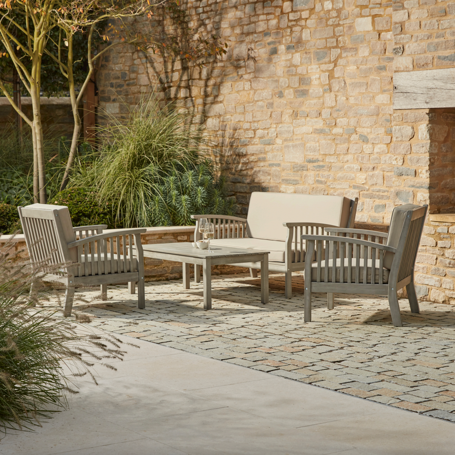 The Repton Wooden Lounge Set, featuring a two-seater sofa, two armchairs, and a matching coffee table in a weathered grey finish. The outdoor seating set includes comfortable beige cushions and is arranged on a stone patio against a rustic brick wall with lush greenery. Designed for stylish and relaxing garden lounging.