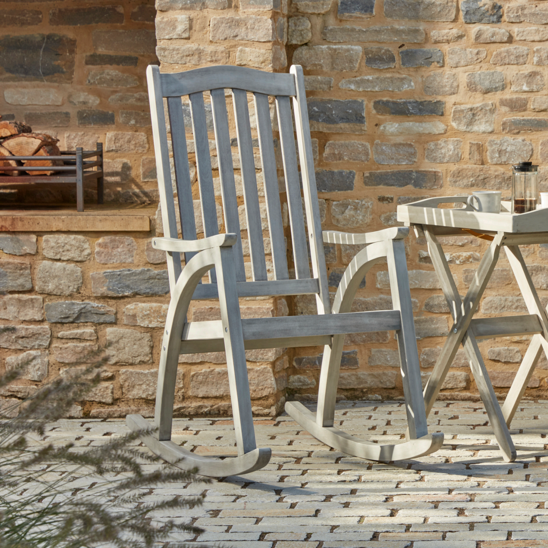 The Repton Wooden Rocking Chair, featuring a classic slatted backrest, curved armrests, and a smooth weathered grey finish. Positioned on a stone patio next to a small wooden side table with a coffee setup, this sturdy outdoor rocker is designed for relaxation in garden or porch settings. Set against a rustic brick wall with a built-in fireplace.