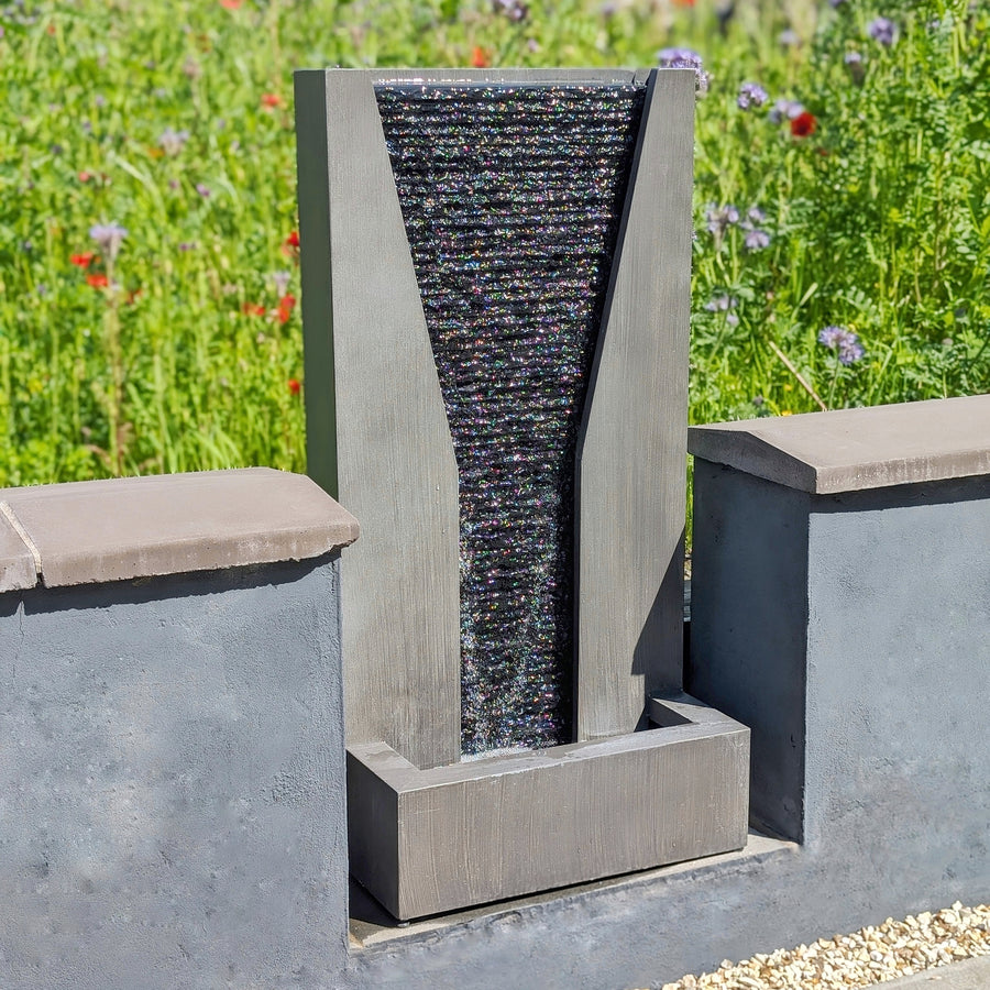 A tall slate water feature with a sleek, modern design, placed in an outdoor setting against a low garden wall, with water cascading down the textured slate surface.