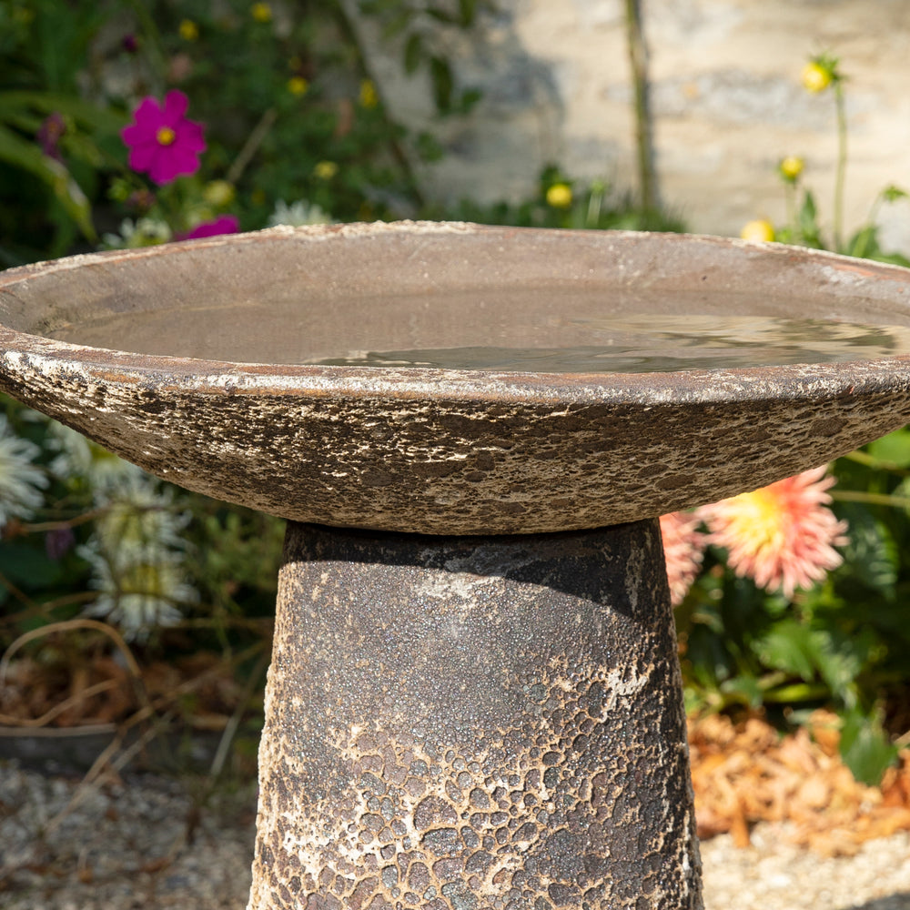 Ancient Salt Glaze Bird Bath nestled among wildflowers, enhancing its organic, timeworn appeal in a natural outdoor space.