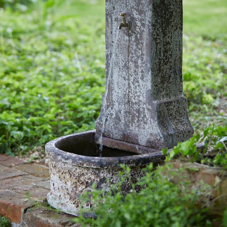 Classic stone water fountain in a garden courtyard, featuring an antique-inspired weathered finish and a traditional standing pillar design.