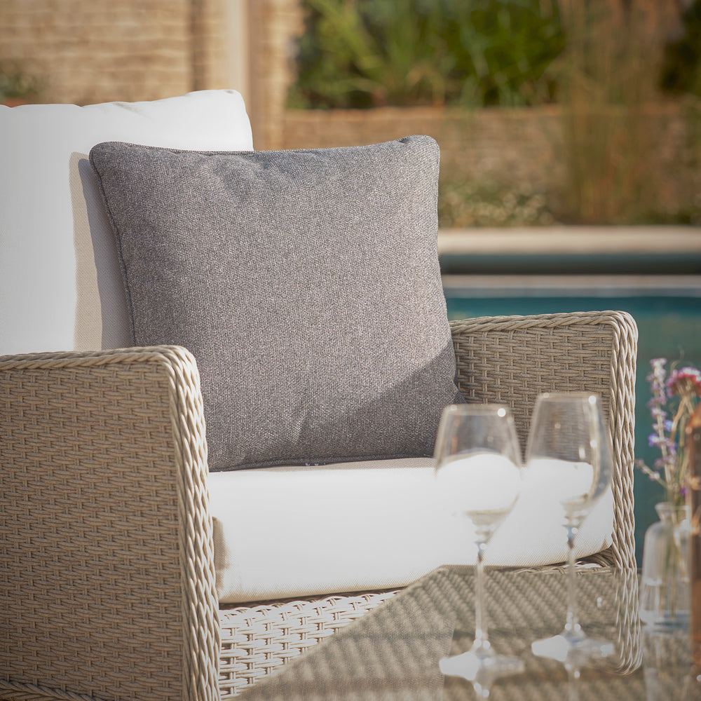 Close-up of the Paxton lounge armchair with soft grey cushions, showcasing elegant woven rattan detailing and a glass of champagne on the armrest.