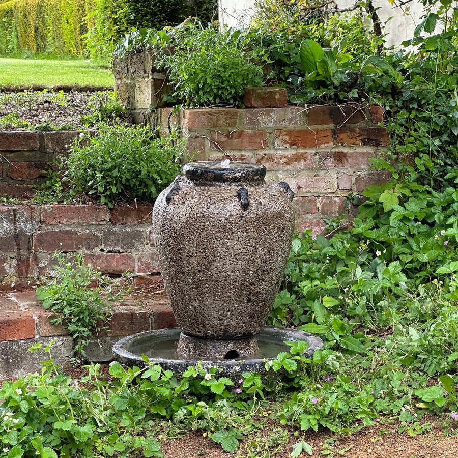 A textured stone water feature with a salt glaze finish, placed in a lush garden setting against a brick wall, surrounded by greenery.