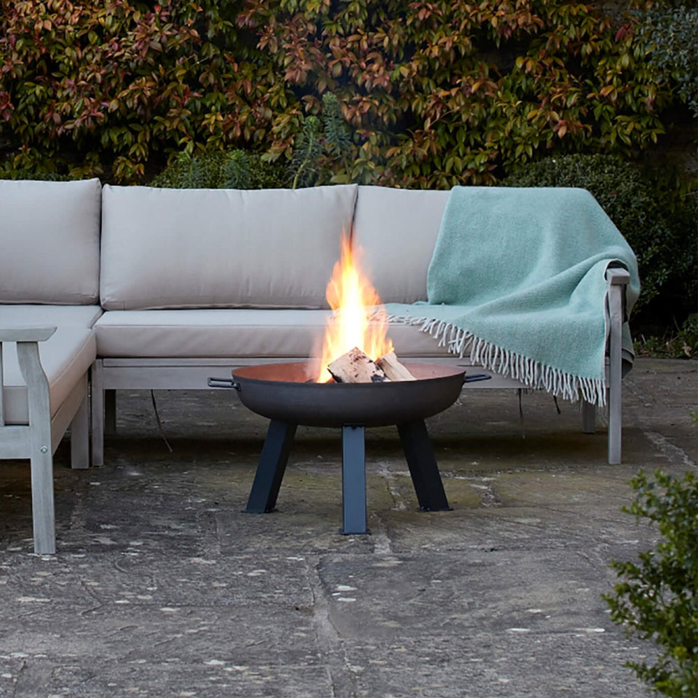 Hoole Cast Iron Fire Pit Bowl in an outdoor seating area, glowing with a fire. A timeless, weather-resistant fire pit designed for cozy garden evenings.