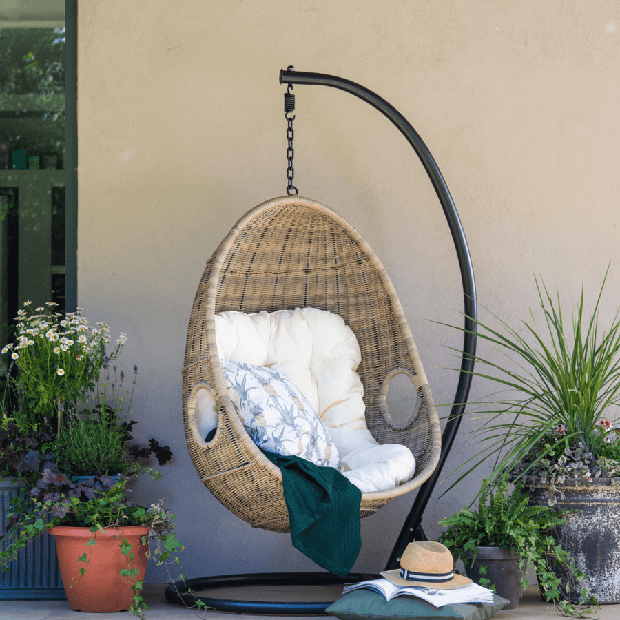 Paxton Rattan Hanging Egg Chair on a patio, styled with plush cushions, a throw, and surrounded by potted plants for a cozy outdoor retreat.