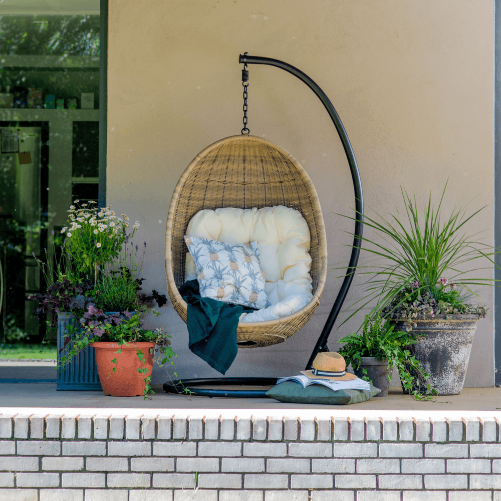 Paxton Rattan Hanging Egg Chair in a neutral-toned garden setting, highlighting its sleek curved frame and woven rattan-effect design.