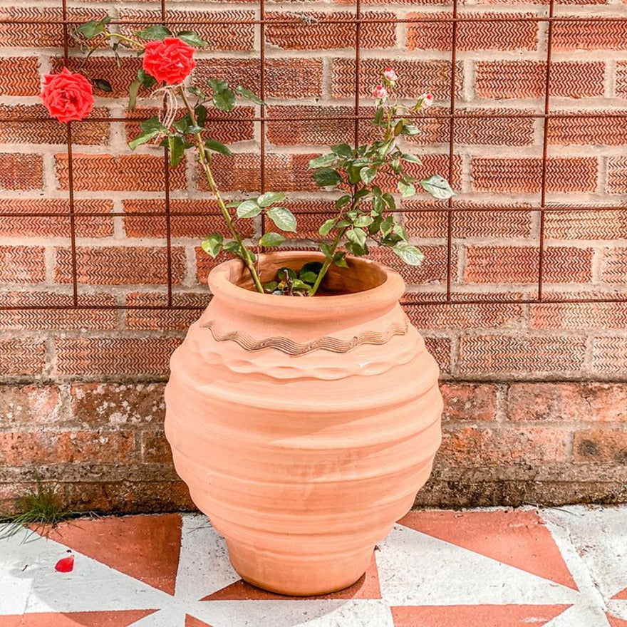 A large Grecian-style terracotta planter with an ornate wavy rim and ribbed texture, placed against a red brick wall, holding a rose plant.