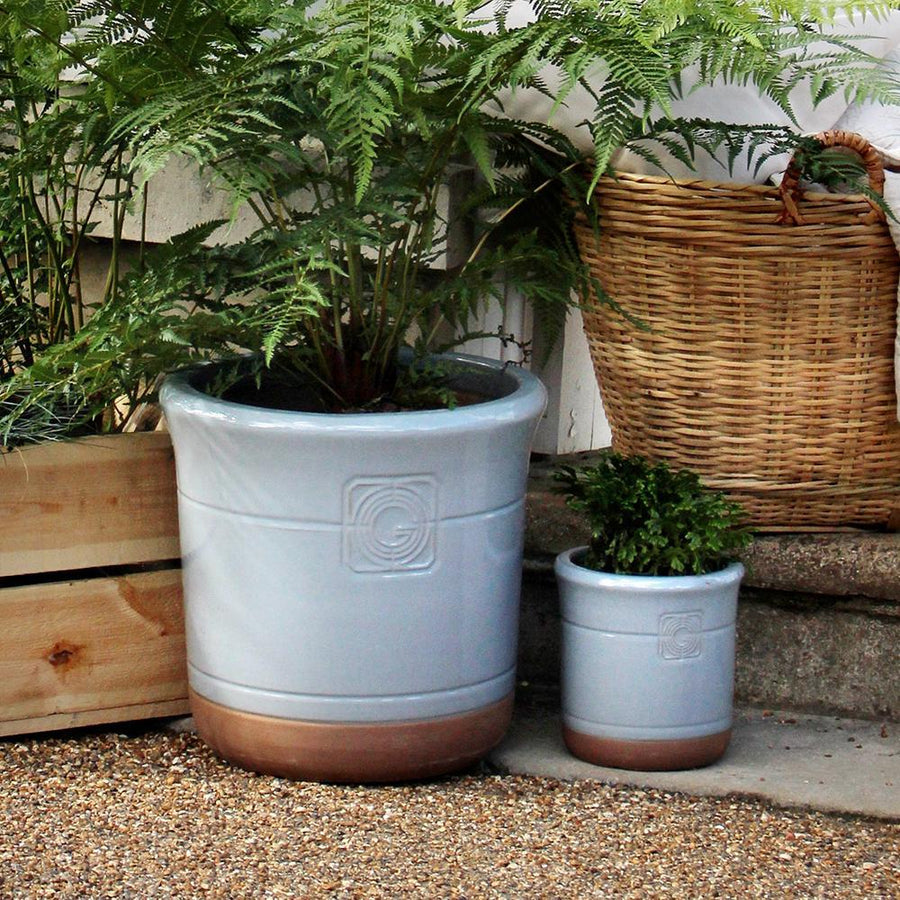 A light blue and terracotta planter with embossed detailing, placed on a gravel surface and filled with lush ferns. A woven basket and wooden planter box sit in the background.