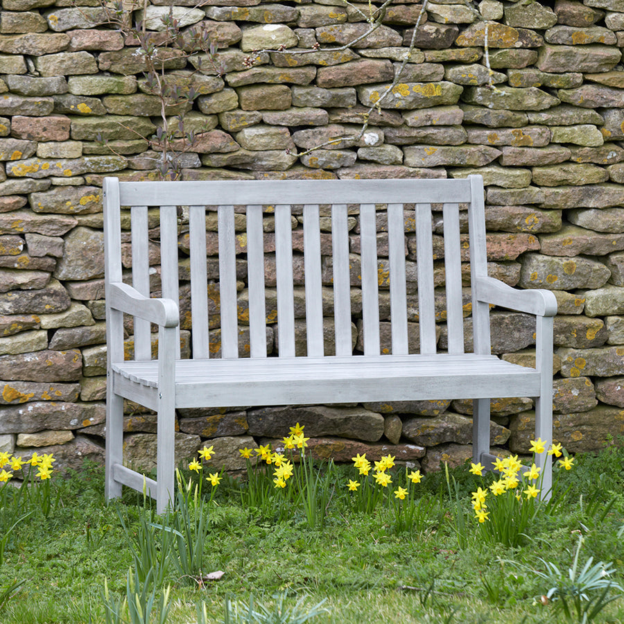 Repton Wooden Garden Bench with a textured grey finish, placed against a rustic stone wall surrounded by blooming yellow flowers. A classic farmhouse-style two-seater bench designed for patios and gardens.