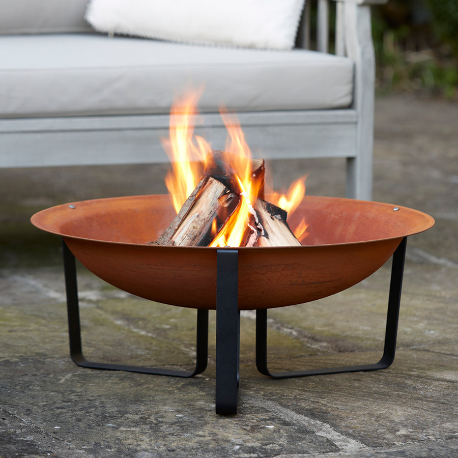 A rust-finish steel fire pit with black metal legs, placed on a stone patio with a fire burning inside, positioned in front of a grey outdoor sofa.