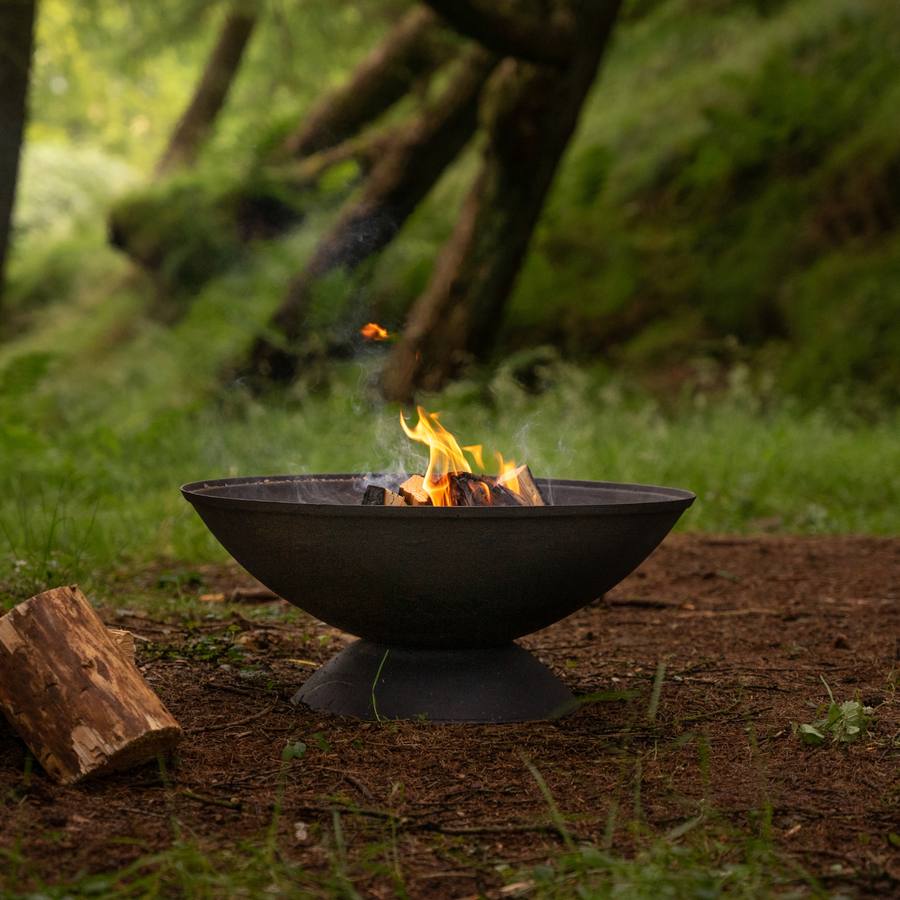 A black cast iron fire pit with a lit fire, placed on a forest floor surrounded by trees, creating a cozy outdoor atmosphere.