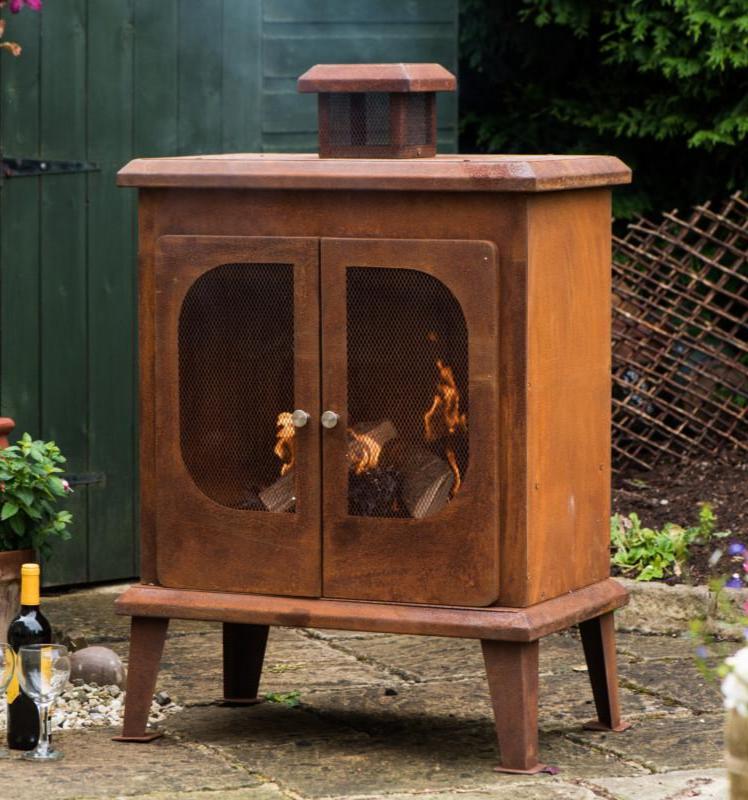 Wakehurst Outdoor Wood Burning Fireplace with a vintage rust finish, placed in a cozy garden setting. A freestanding steel log burner with mesh doors and a built-in chimney for efficient smoke control.