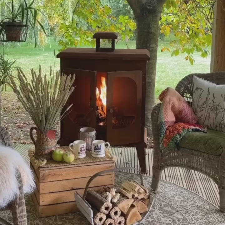 A garden scene showcasing the Wakehurst Outdoor Fireplace in use, with a glowing fire and a cozy outdoor seating setup. Watch the video to see this stylish steel log burner in action.